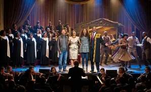 A choir sings on stage with a cityscape backdrop and red velvet curtains.