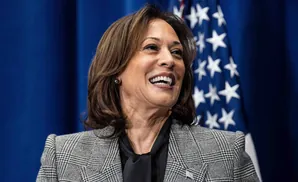 Kamala Harris smiles in a grey plaid jacket and black shirt in front of an American flag.
