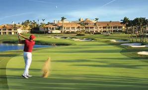 Golfer in red shirt and white pants swings club on a golf course with sand flying.