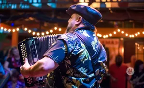 Man in a floral shirt and cap plays an accordion at an outdoor concert with string lights.