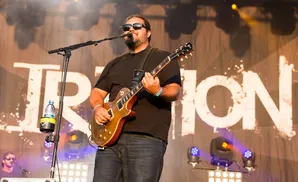 A guitarist with sunglasses and a beard plays an electric guitar on stage.