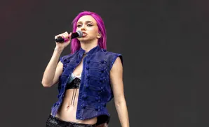 A woman with bright pink hair sings into a microphone while wearing a blue denim vest over a sparkly top and black skirt.