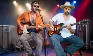 Two men playing guitars on a stage, one singing into a microphone, under colorful stage lights.