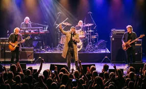 A band performs on stage, with a singer holding a flute and other musicians playing guitar, bass, and keyboards to an audience.