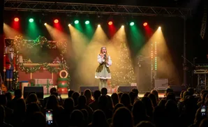 A singer dressed as an elf performs on a stage decorated for Christmas, with a live audience watching.