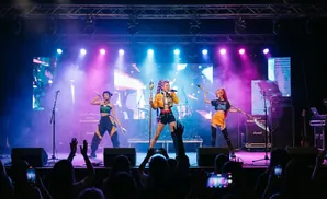 Three women in cosplay outfits perform on stage with purple and blue lighting.