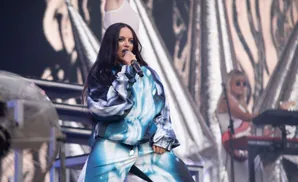 A woman with long dark hair sings into a microphone while wearing a blue and white patterned tracksuit on stage.