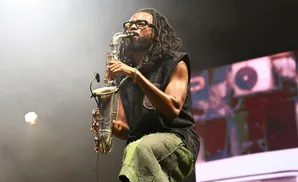 A man with dreadlocks plays a saxophone onstage, wearing sunglasses and green pants.