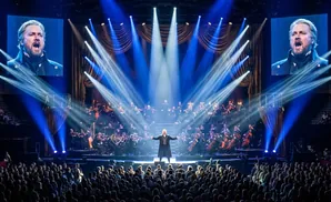 A cast of singers in period costumes performs on a dimly lit stage with dramatic red and blue lighting.