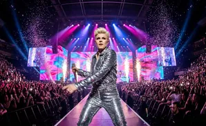 A singer in a sparkling silver outfit performs on stage with confetti falling and the audience cheering.