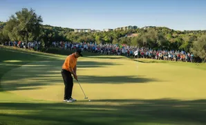 Golfer in orange shirt putts on a green with a large crowd watching from the hillside.