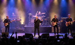 A band performs on stage with a singer in the front, guitarists and a keyboardist behind, and a drummer in the back.