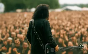 A musician with long, dark curly hair wearing a black studded shirt plays a black bass guitar on a stage in front of a blurred, cheering crowd.