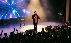 A comedian performs for a cheering audience on a dimly lit stage with a colorful screen behind him.