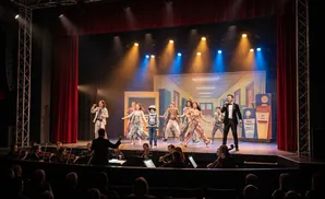 Stage performers in costumes dance and sing on stage with an orchestra in the foreground.