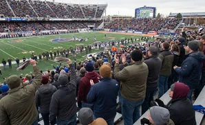 Fans wave towels at a football game, with a packed stadium and scoreboard visible in the background.