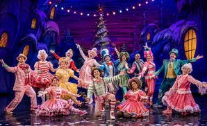A group of performers in whimsical, colorful costumes on a stage with a fantastical, cartoonish backdrop.