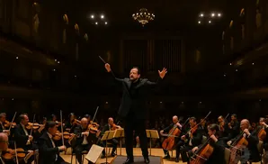 A conductor leads an orchestra with violins and cellos, bathed in warm stage lights.