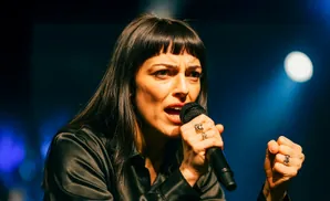 A woman with bangs and long dark hair sings into a microphone, wearing a black shirt and rings on her fingers.
