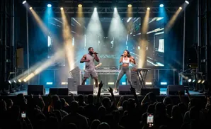 Performers sing and dance on a stage with bright lights and screens, to an audience with their phones up.