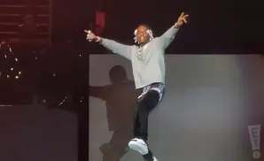 A Black man with white braided hair, wearing a gray sweatshirt and black pants, dances on stage with his arms raised.