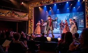 A stage play with actors in Victorian costumes performs a Christmas scene in front of a snowy London backdrop.