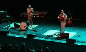 Musicians perform on a dimly lit stage, with a drummer on the left and a guitarist on the right.
