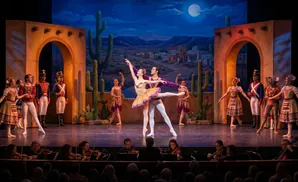Ballet dancers perform on stage with a desert backdrop and orchestra in the foreground.