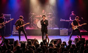 A male singer and band perform live on stage to an enthusiastic audience, bathed in purple and blue stage lights.