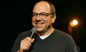 A smiling man with glasses holds a microphone to his mouth, with a dark background and blurred lights.