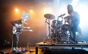 Two musicians in black and white polka dot outfits perform on stage. One plays a double-neck guitar, the other a drum kit.