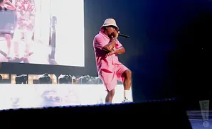 A rapper in a pink bucket hat and matching pink shorts suit performs on stage, holding a microphone.