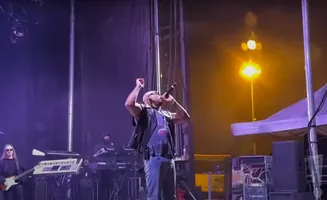 A bald male singer with a shaved head performs on stage, holding a microphone to his mouth, wearing a black vest over a graphic t-shirt and blue jeans.