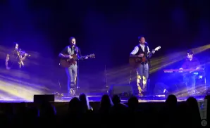 A band performs on stage with blue lighting, featuring a violinist, two guitarists, and a keyboardist.