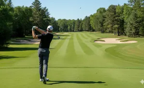 Golfer in black shirt and gray pants swings at a golf ball on a sunny day at a course with sand traps.