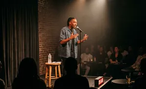 A Black comedian performs on stage in front of a brick wall while an audience watches.