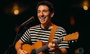 A smiling man in a striped shirt plays an acoustic guitar and sings into a microphone on stage.