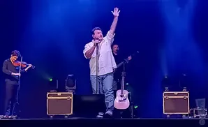 A male singer on stage with his arm raised, holding a microphone. A violinist stands to his left, and a guitarist is behind him.