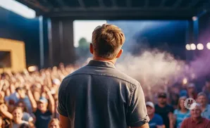 Man with blonde hair on stage, facing a large cheering crowd at an outdoor concert.