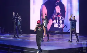 A group of five male singers perform on stage under bright lights, with a large screen behind them displaying a band member.