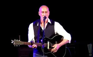 A bald man in a white shirt and vest plays an acoustic guitar and sings into a microphone.
