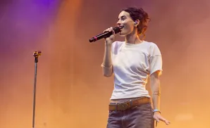 Singer on stage holds a microphone, wearing a white shirt, jeans, and a brown belt with an ornate buckle.