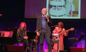A man holds a saxophone while standing center stage; a woman sits left playing piano, and a man plays bass guitar right.