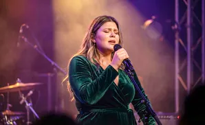 Woman in green dress sings into a microphone on stage with purple and orange lighting.