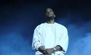 Kanye West in a white sweatshirt and gold chain on stage with blue smoke.