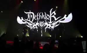 A band performs on stage with a large white Dethklok logo behind them.