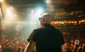 Bald man in black t-shirt and sunglasses on stage facing a cheering crowd under bright spotlights.