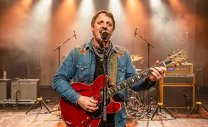 Man in a denim shirt with embroidered flowers plays a red guitar on stage with lights and smoke.