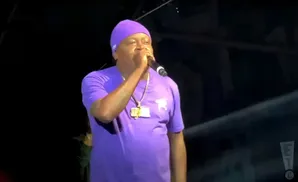 A man in a purple t-shirt and beanie holds a microphone, performing on stage.