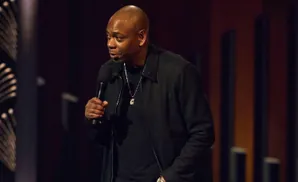 Dave Chappelle on stage holding a microphone and wearing a black jacket and necklace.